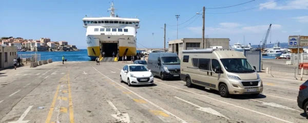 File de véhicules de différentes tailles — citadine blanche, fourgon gris et camping-car beige — attendant l'embarquement sur un quai de ferry méditerranéen sous le soleil estival